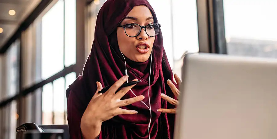 Una donna che indossa occhiali da lettura e un hijab viola gesticola mentre parla guardando un computer portatile o uno schermo desktop, potenzialmente in una call.